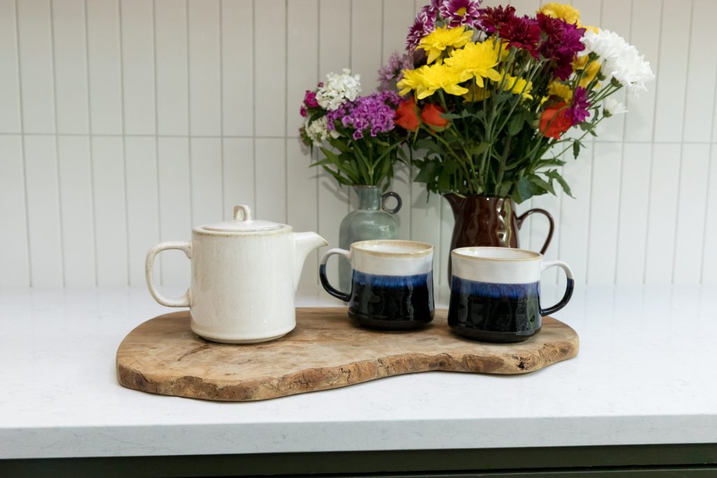 Cream teapot and blue reactive glaze mugs on a wooden board with flowers