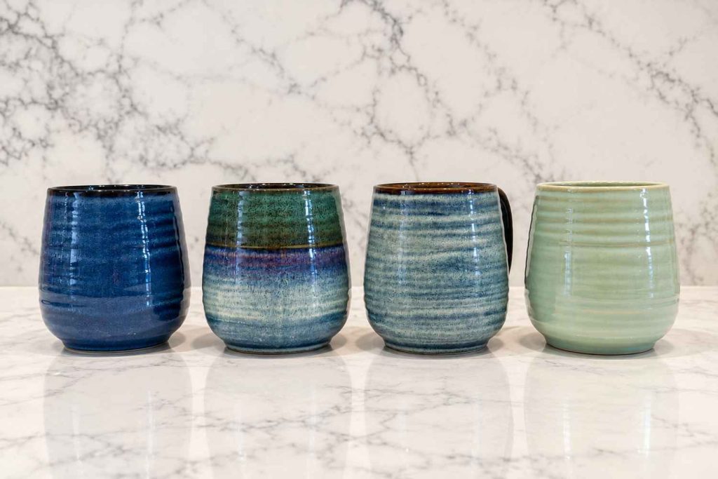 Four reactive glaze ceramic mugs in navy, blue, grey and sage lined up on a marble worktop